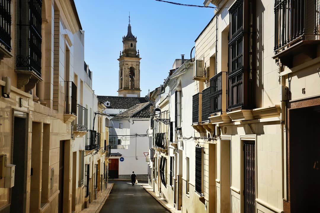 Calles de Gilena conduciendo hacia el campanario de la iglesia, destacado bajo un cielo azul claro.