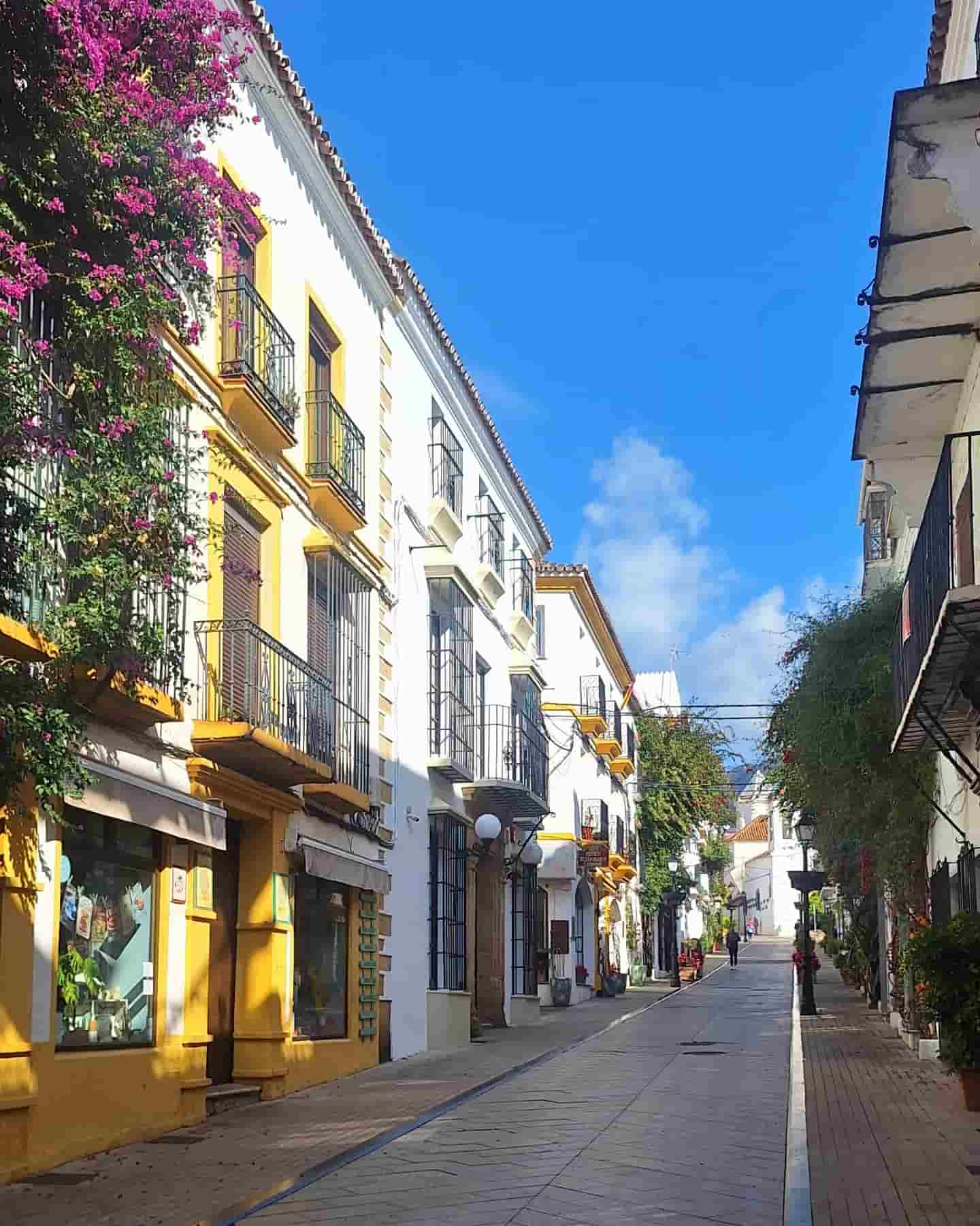 Calle céntrica de Marbella con balcones soleados y adornados con flores coloridas.